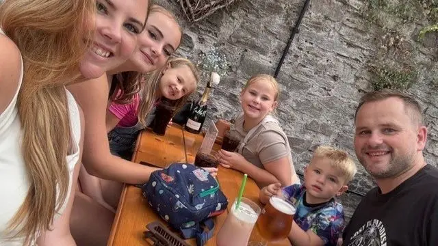 A family is shown smiling and seated around a wooden picnic table as the mother reaches out her hand to take a selfie of the group.