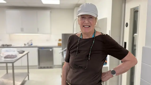 A woman stands in an industrial looking kitchen. She has on tan trousers, a brown top and a grey baseball cap.