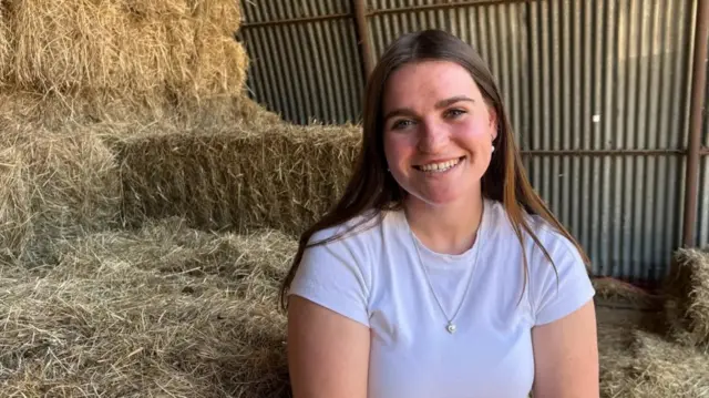 Lydia Law looking directly to camera and smiling. She is wearing a white, round-necked T-shirt and has straight brown hair, worn down and reaching over her shoulders. She is seated in a hay barn, with bales of hay seen behind her on the left of frame and a corrugated metal wall in the further distance.