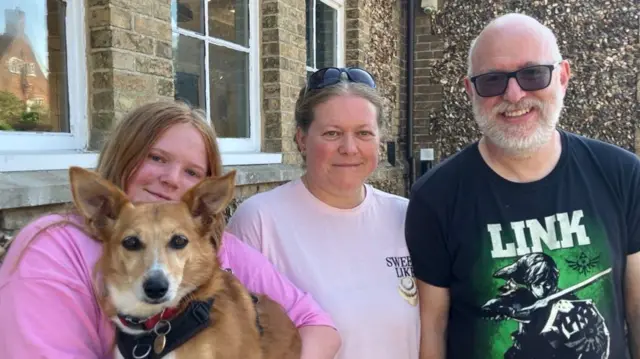 A young woman holding a dog stands beside her mother and father who are all smiling at the camera. The young woman has long red hair and is wearing a pink top. The mother has tied up blonde hair and wears a lighter pink T-shirt with sunglasses on her head. The man is bald with a grey beard, sunglasses and a black T-shirt.
