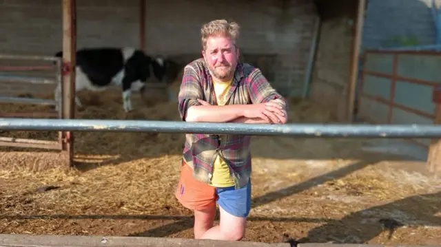 Nicholas Millard - a man with blonde hair, beard and reddish face leans on a metal railing in a checked shirt, yellow T-shirt and shorts, one leg orange and the other blue. Behind him is hay and a cow stands in the background to his left.