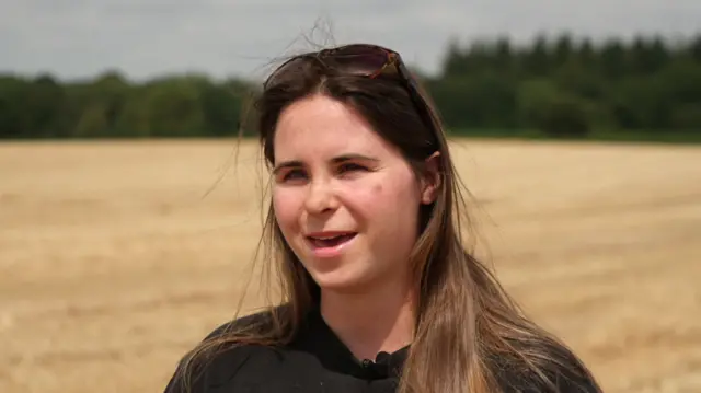 Eleanor Gilbert stands in a wheat field. She has long brown hair and is wearing a block top.