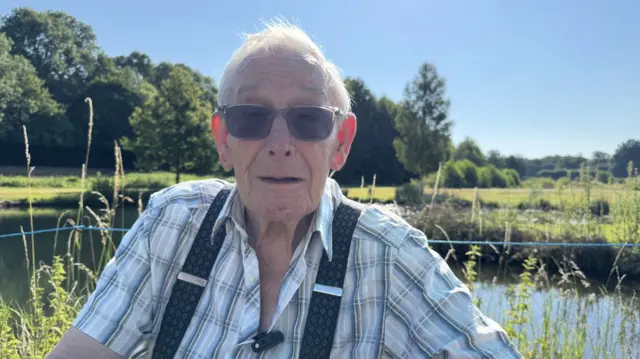 Barrie Arminson stands in front of a river, wearing sunglasses, a checked short-sleeved shirt and braces.