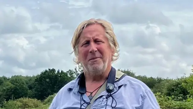 A man wearing a blue shirt and holding a pair of binoculars which are hanging around his neck. He is standing in a woodland with trees and bushes in the background.