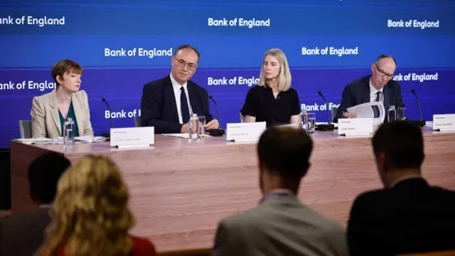 Bank of England news conference, showing four people sitting behind a panelled wooden desk in front of a blue background. The backs of the heads of several journalists are visible in the foreground.
