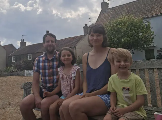 Man and a woman with brown hair in summer clothes sitting next to two young children, a girl and a boy.