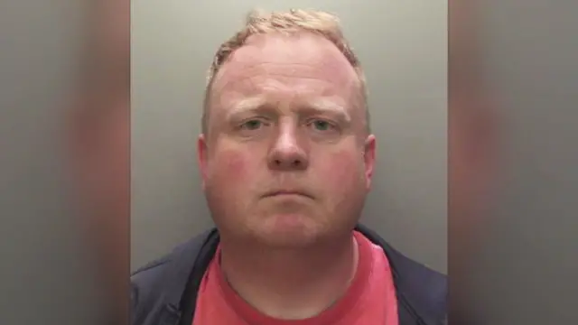 Police custody picture of Robert Bush he has with short, light brown hair is and is wearing a dark jacket over a red shirt.