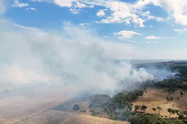 Plumes of smoke billow over dry brown fields and green trees. The white dome of a power station is visible several miles further south, in the background.