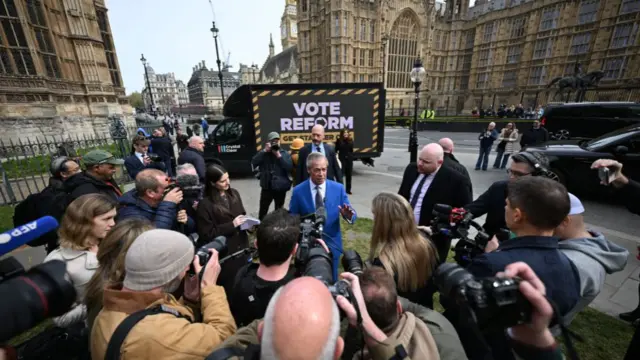 Nigel Farage, wearing a blue suit, speaks to a semi-circle of journalists pointing cameras at him. He is standing in front of a billboard outside the Houses of Parliament