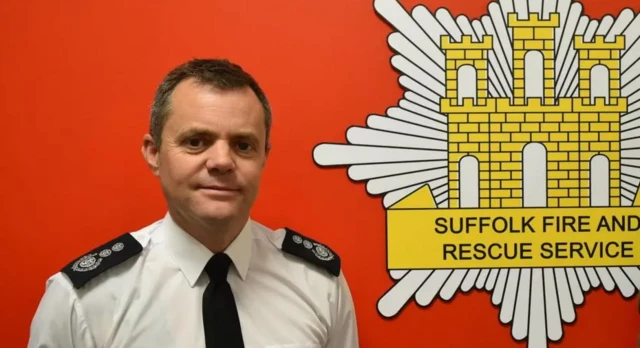 Jon Lacey stands in front of a red background with the yellow Suffolk fire and rescue badge on it. He is wearing a white shirt and black tie.