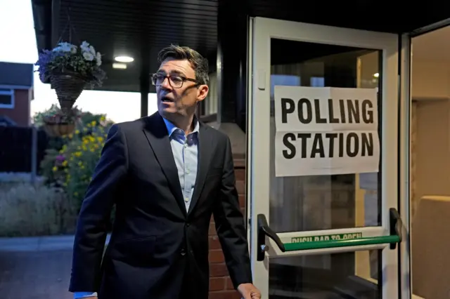 Andy Burnham wearing a dark suit pictured walking through open doorway. There's a large paper sign stuck to the clear door reading: "Polling Station"