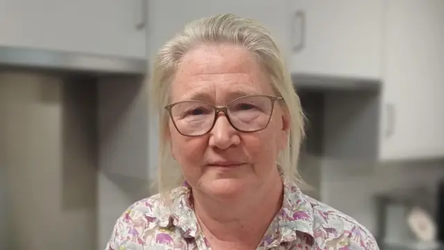A woman stands inside a property. She is slightly smiling at the camera. She has grey hair that has been tied back and she is wearing a colourful shirt with various different animals printed on it.