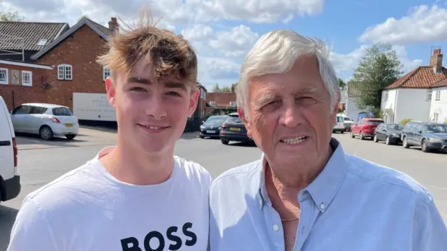 A teenage boy wearing a white T-shirt and who has brown hair stands in a village alongside his granddad. The older man has grey hair and is wearing a blue shirt. Parked cars and homes are behind them.