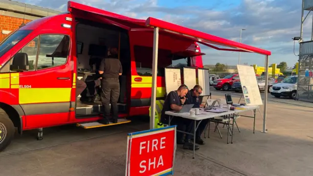 A fire service vehicle parked in a car park. Its side doors have been opened up with a desk sat outside where two firefighters sit and work.