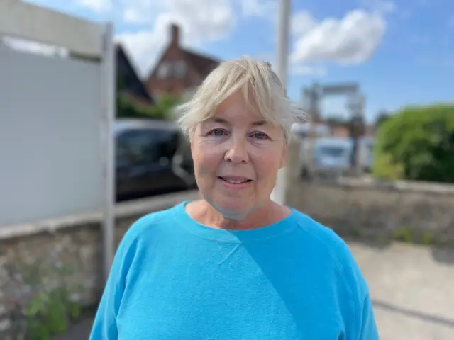 Jane Thompson wears a blue T-shirt. She has blonde hair and looks at the camera, standing outside.