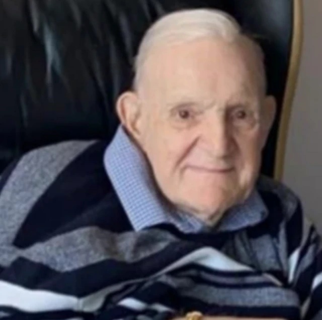 An older man is shown smiling and hunched in a chair, wearing a striped blue top and shirt.