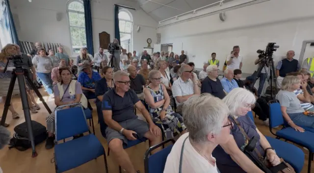 People sitting on chairs in rows during a meeting at a village hall. They are all facing toward the right which is the front of the hall