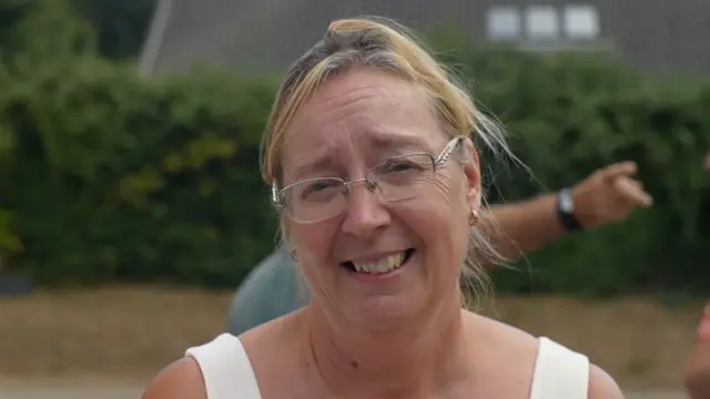 A woman smiles at the camera as she stands outside. She has blonde hair that is tied back, glasses and she is wearing a white vest top. Some other people are behind her in the background.