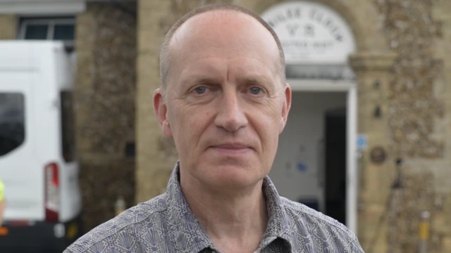 A man stands outside a village hall. He is slightly smiling at the camera. He has shaved hair and is wearing a grey shirt.