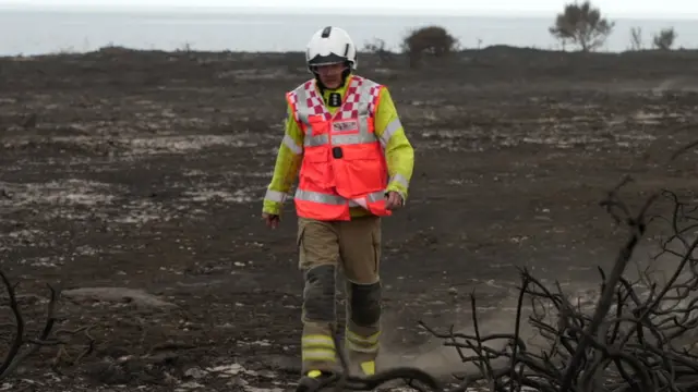 A firefighter with his head down walking across a burnt landscape at Dunwich Heath. He is wearing a white helmet and orange hi-vis over a yellow jacket.