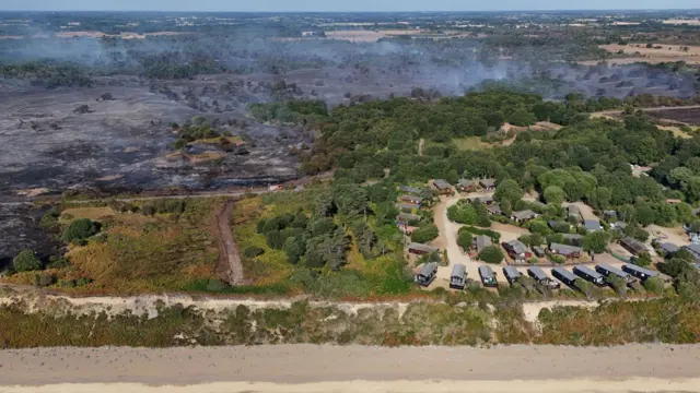 A drone image above a coastline where fields have been scorched black during a fire. A nearby holiday park with caravans can be seen on the right that the fire had not reached.
