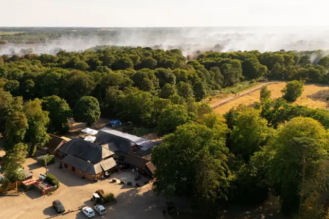 A farm house and parked cars are in the foreground. Behind are green trees, and further back, white smoke smouldering from the ground.