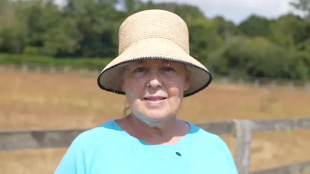 A woman smiles at the camera as she stands in a field. She wears a cream hat and blue T-shirt.