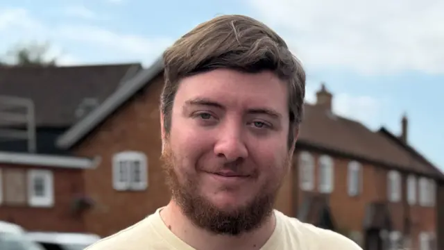 A man stands on a residential road smiling at the camera. He has brown hair which has been swept across one side of his head. He has a brown coloured beard and is wearing a yellow T-shirt.