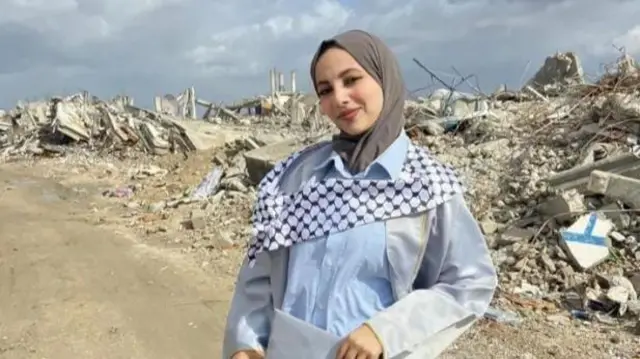 Tala stands holding graduation cap in front of debris from warzone
