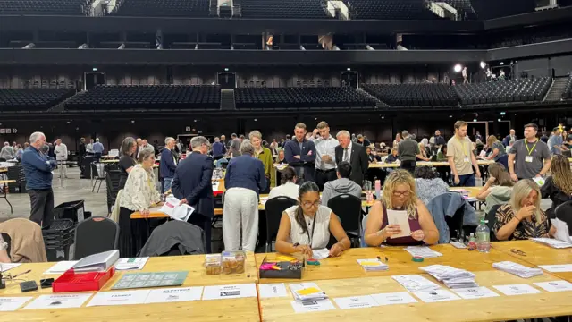 People sitting at counting tables in an arena.