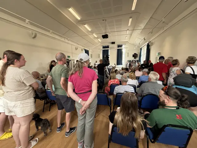 The backs of people sitting and standing in a village hall.