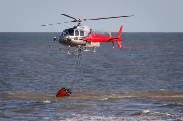 A red and silver helicopter hovering over the North Sea at Dunwich. It is trailing a large red bucket into the sea, collecting water.