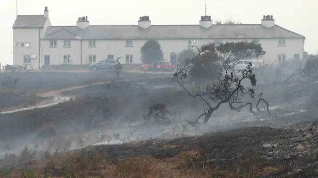 A long terrace of white cottages called Coastguard Cottages seen through the smoke. In front of them, there are burnt out shrubs and a burnt landscape, which is smoking.