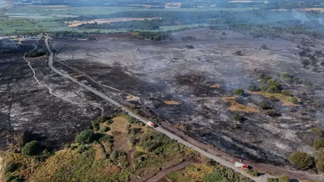 A drone image above heathland that has been scorched black during a wildfire. Smoke billows up from the ground. There is a road through part of the area where fire engines drive along.