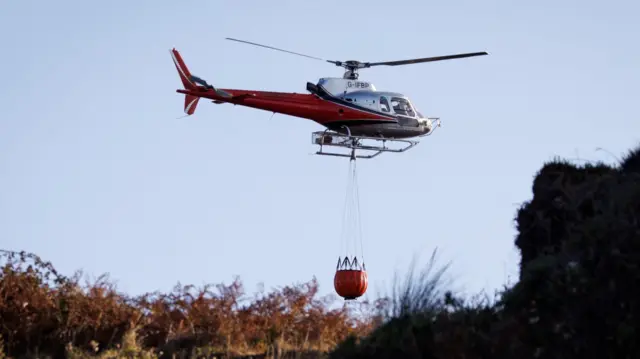 A red and silver helicopter flying over heathland with a red bucket of water underneath.