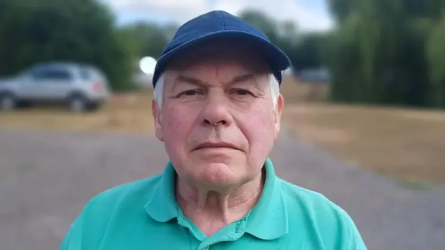 A man stands outside in a car parking area on a camping site. He wears a blue cap with a light green polo top. He is slightly smiling as he looks directly at the camera and some grey hair can be seen under his cap. The background behind him is blurred out of focus.