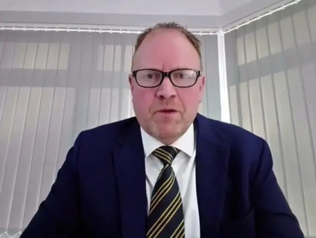 A man wearing a dark suit and a stripy tie is pictured in front of vertical blinds