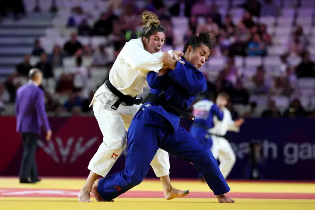 Acelya Toprak, wearing a white robe, and Lele Nairne, wearing a blue robe, both of Team England, fight in the women's 57kg semi-final