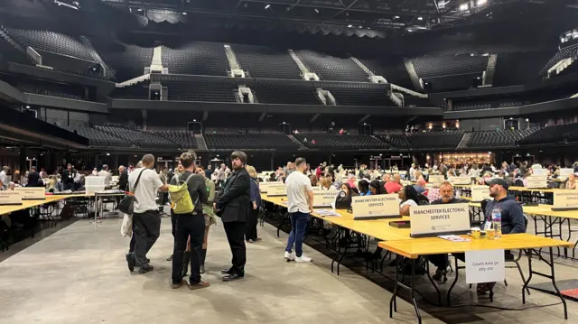 Rows of tables with people sitting at them, while others stand on the floor of the arena.