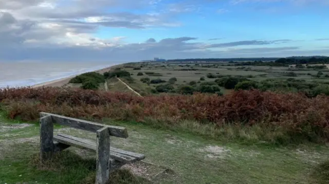 A wooden bench is in the corner of the frame with a view of the coastline to the left and the heath to the right