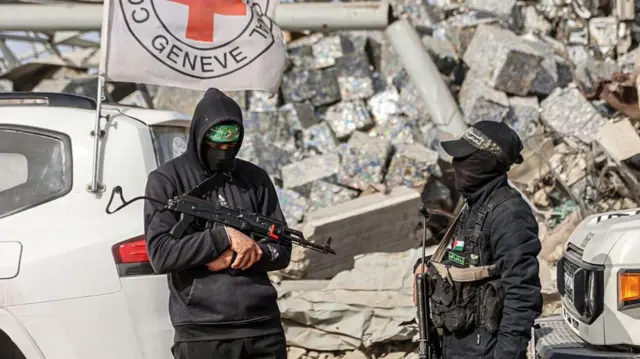 Two men with firearms dressed head to toe in black are pictured in front of Red Cross vehicles and a pile of rubble.