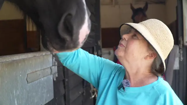Jane wearing a blue top and cream hat, looks up to a horse at a stable, and brushes its mane.