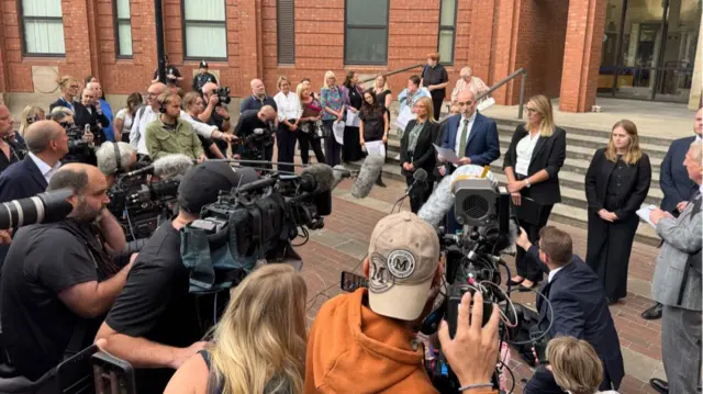 A crowd is gathered around people speaking behind microphones outside court.