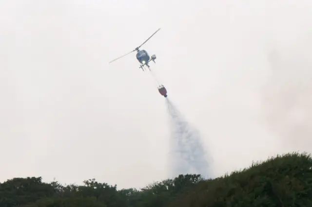 A helicopter flies water collected from the sea to drop on the wildfire at Dunwich Heat. The water is being dropped.