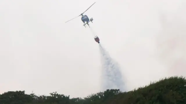 A helicopter is shown dropping water over heathland hills against a smoky sky.