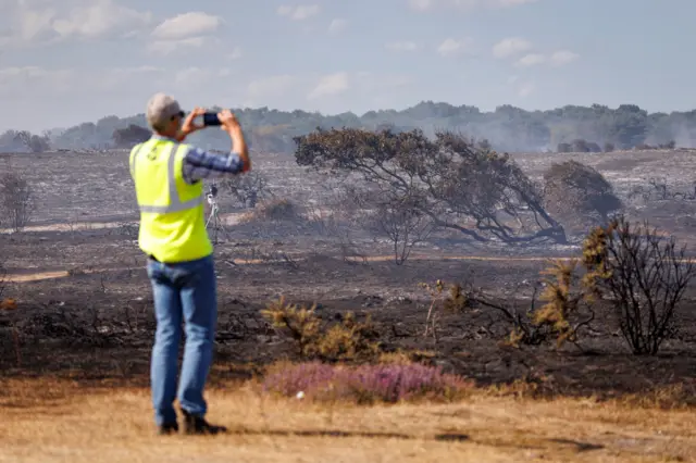 A National Trust worker in a hi-vis yellow jacket monitoring the fire at Dunwich Heath. He is standing on an unburnt patch of yellow grass and beyond him is blackened landscape, from which smoke rises.
