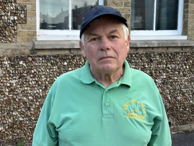 David Watson in a turqoise jumper and a navy cap. He is standing in front of the village hall.