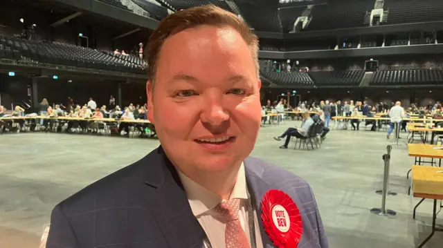 Labour MP for Stretford and Urmston, Andrew Western, has short brown hair and is wearing a navy suit with a pink tie