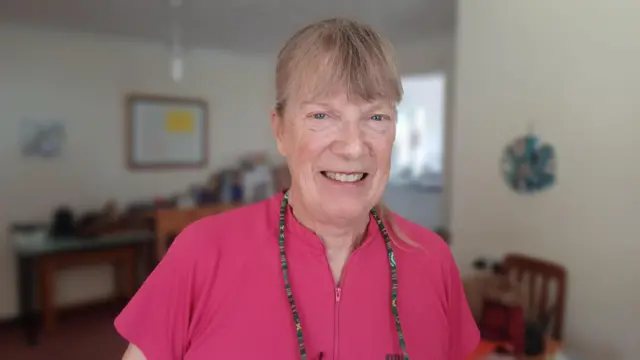 A woman stands inside a home and smiles at the camera. She has light brown hair with some grey in it that has been tied back. She is wearing a pink zip up top with glasses that hang on a fabric chain around her neck.