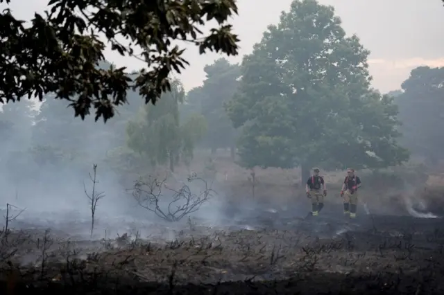 Firefighters walk through the smouldering remains of a wildfire at Dunwich Heath. Smoke continues to rise around them and everything else is black.